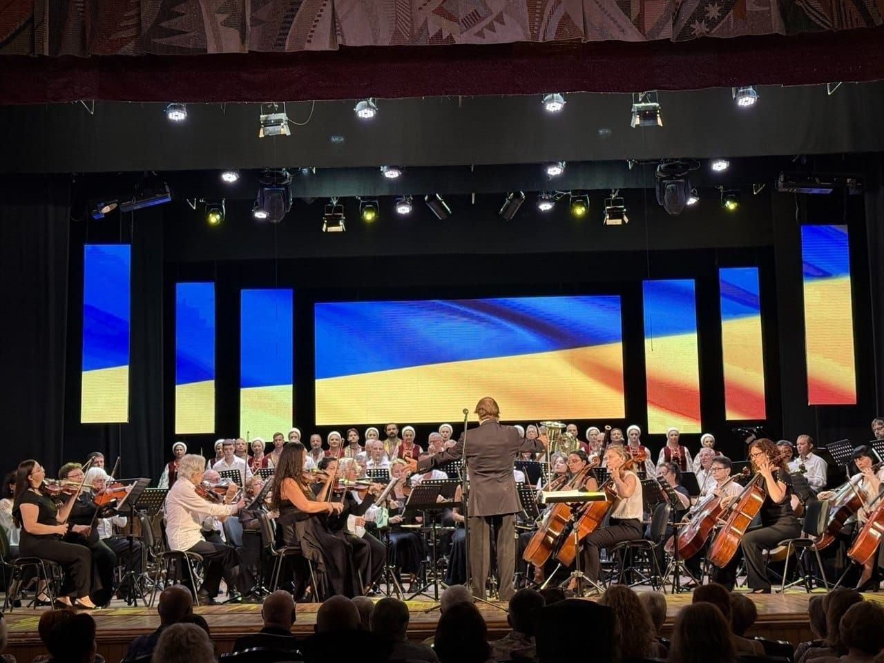 У Черкаській обласній філармонії ім. Олександра Кошиця відбувся урочистий концерт «З молитвою за Україну» до Дня Незалежності України, організований для переселенців з Херсонщини за сприяння Чаплинської та Долматівської СВА. На фото головний диригент Київської опери керує великим симфонічним оркестром та народним хором на сцені філармонії на тлі світлодіодних екранів із зображенням синьо-жовтого державного прапора України.