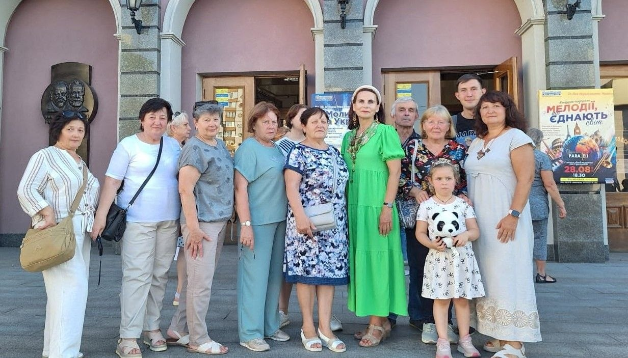 Група переселенців з Херсонщини (дорослі та маленька дівчинка з іграшковою пандою) стоїть разом перед входом до будівлі Черкаської обласної філармонії ім. Олександра Кошиця після урочистого концерту «З молитвою за Україну» до Дня Незалежності України, організованого Чаплинською та Долматівською СВА. На задньому плані видно дерев'яні двері філармонії, барельєф та святкові афіші, зокрема плакат концерту «Мелодії, що єднають світ».