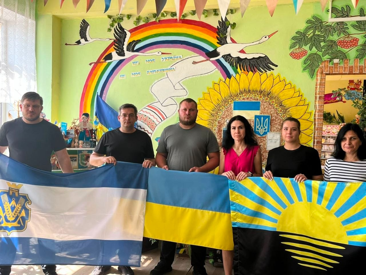 З нагоди Дня Державного Прапора України та Дня Незалежності України представники Координаційного центру «Вільні разом» у Кривому Розі долучилися до патріотичного флешмобу «В єдності наша сила» спільно з командою осередку підтримки мешканців Бахмутської громади «З Бахмутом у серці». На фото три чоловіки та три жінки тримають разом три прапори: ліворуч — прапор Херсонської області (із білою та синіми смугами й гербом), по центру — державний прапор України, а праворуч — прапор Донецької області (із чорним низом та золотим сонцем, що сходить на синьому тлі). Захід проходить у просторі на тлі дитячого патріотичного малюнка з соняшником, лелеками та веселкою. Під час цієї зустрічі команди також обговорили плани щодо подальшої співпраці та організації спільних заходів для всебічної підтримки внутрішньо переміщених осіб з Херсонщини та Донеччини.