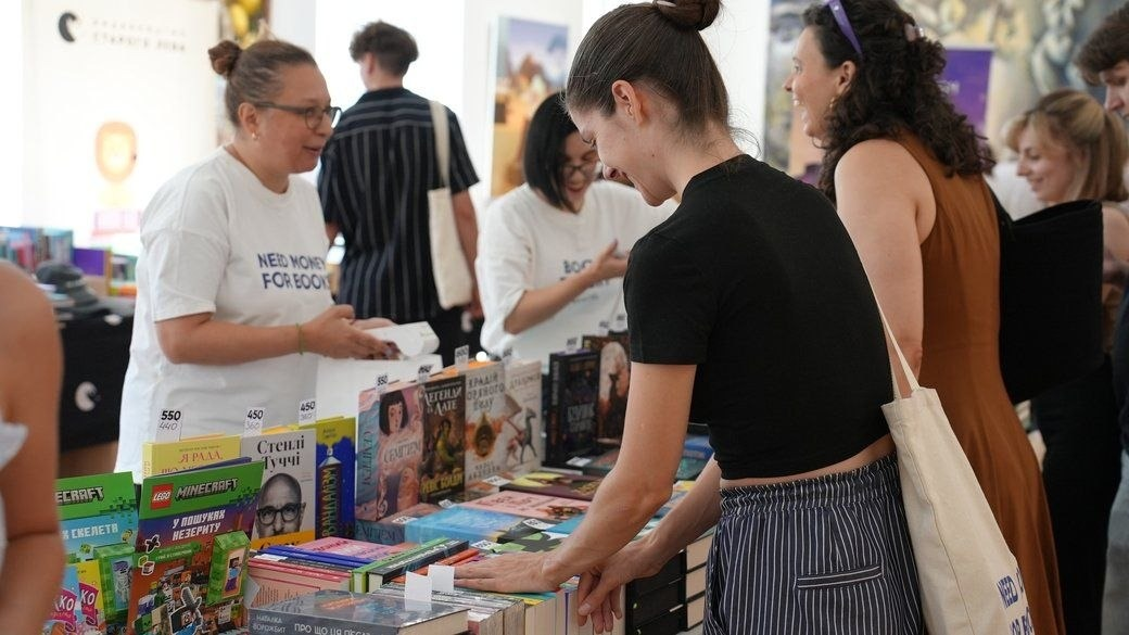 У межах святкових заходів до Дня Незалежності України у Черкасах, організованих за підтримки Чаплинської та Долматівської СВА, переселенці з Херсонщини відвідали літературний фестиваль «Змістовно». На фото зафіксовано ярмарок видавництва «Видавництво Старого Лева», де відвідувачі розглядають та купують нові книжки на розкладках, спілкуючись із консультантками у фірмових білих футболках із написом «NEED MONEY FOR BOOKS». Попереду на столах представлено різноманітну художню та дитячу літературу (зокрема серії Minecraft, книжки Стенлі Туччі).