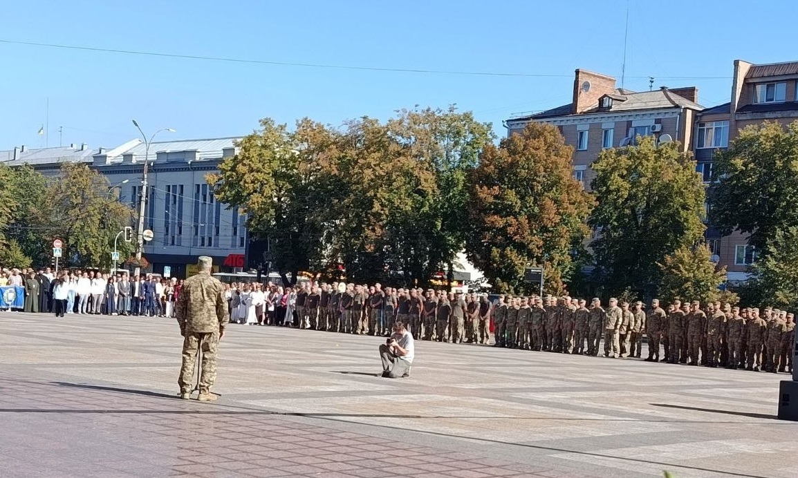 У межах урочистостей з нагоди Дня Незалежності України, організованих Черкаською ОДА за підтримки Чаплинської та Долматівської СВА, переселенці з Херсонщини долучилися до патріотичного заходу просто неба. На фото зафіксовано велику міську площу, де перед військовим керівником довгою шеренгою вишикувалися українські захисники для церемонії нагородження воїнів та родин загиблих героїв. Ліворуч на площі зібралися цивільні мешканці, ВПО, представники духовенства та громадськість на тлі міської архітектури й зелених дерев.