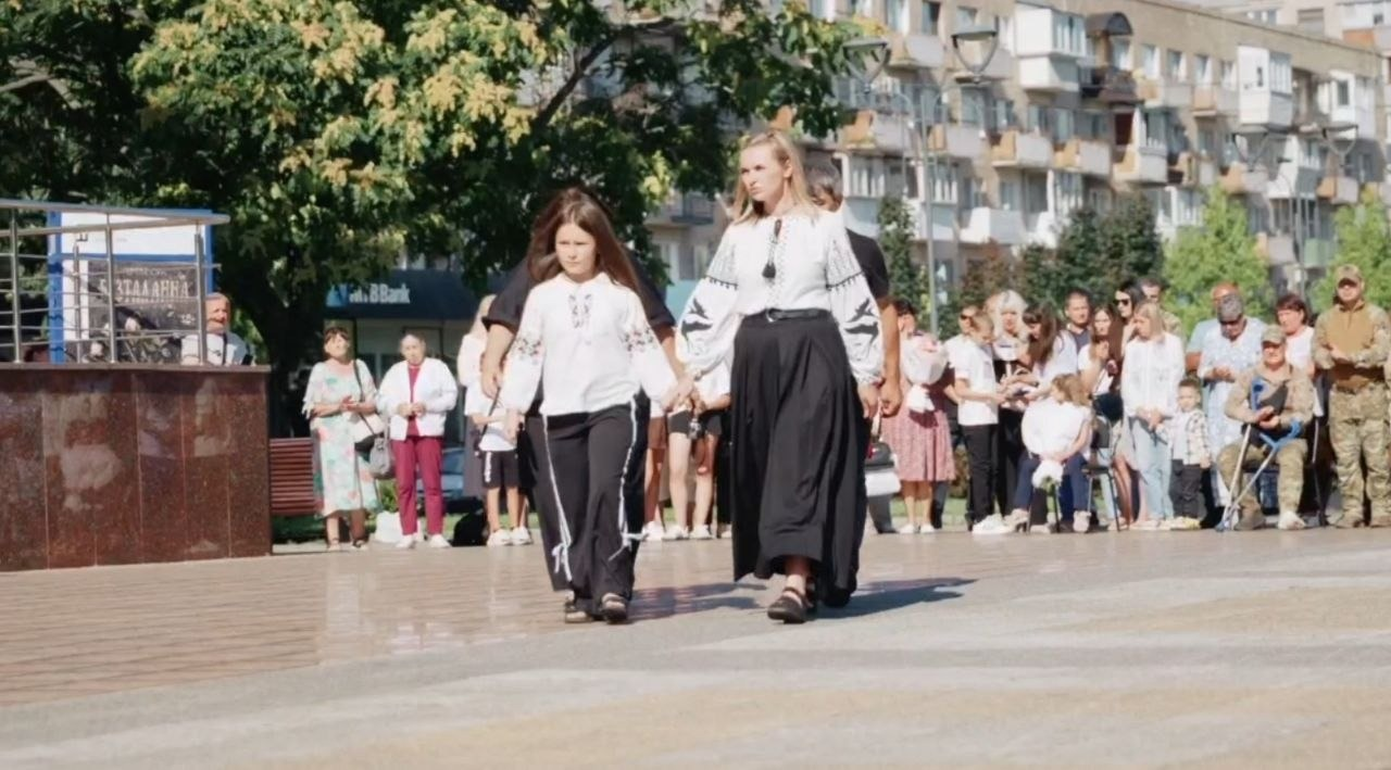 У межах урочистостей з нагоди Дня Незалежності України, організованих Черкаською ОДА за підтримки Чаплинської та Долматівської СВА, переселенці з Херсонщини долучилися до патріотичного заходу просто неба. На фото зафіксовано жінку та дівчинку у традиційних українських вишиванках і чорному вбранні, які йдуть міською площею під час виступу чи церемонії нагородження. На задньому плані за ними спостерігають численні глядачі, серед яких присутні українські військові, цивільні мешканці, ВПО та діти на тлі міської архітектури й зелених дерев.