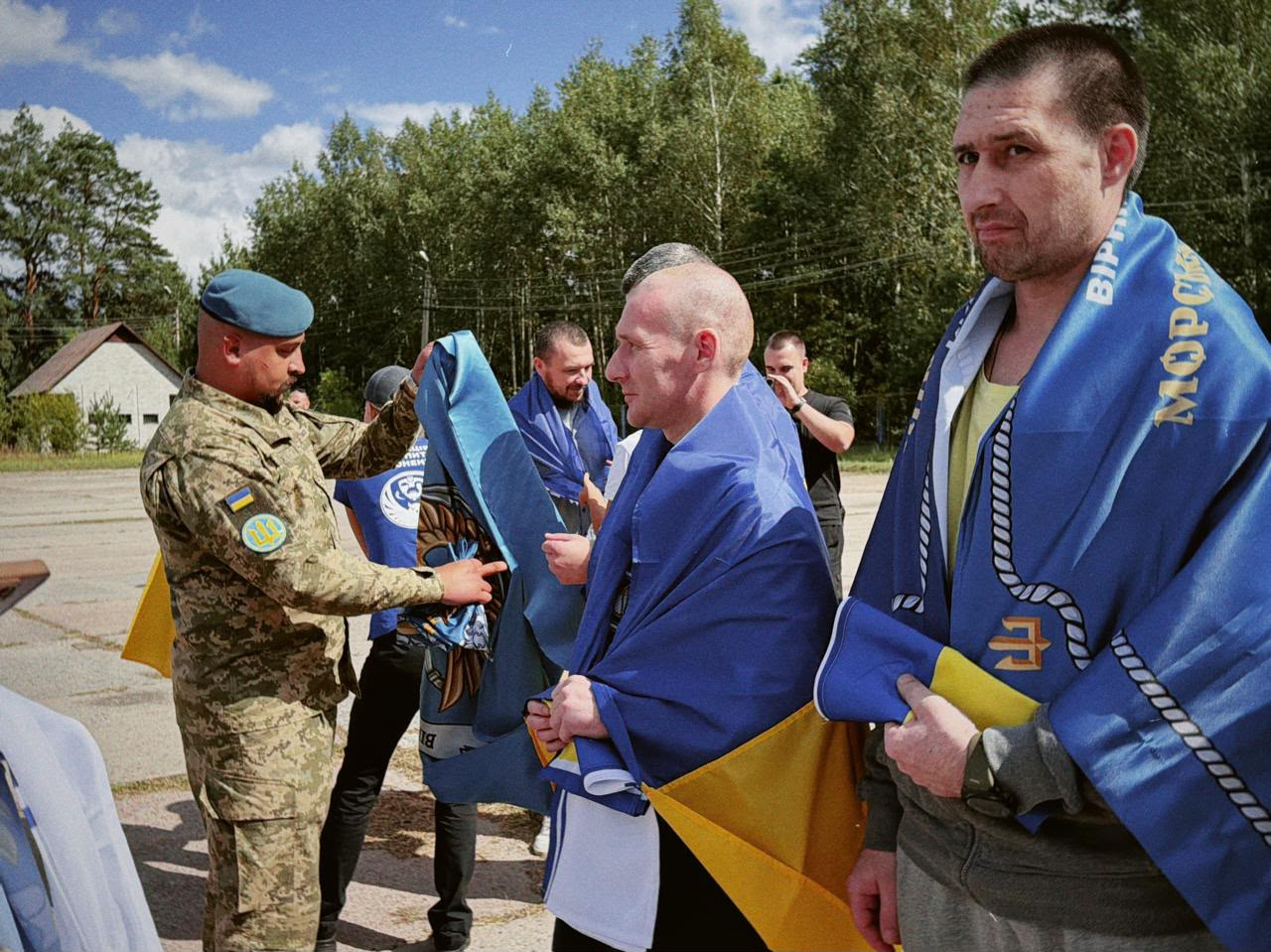 У межах спеціального етапу обміну через територію Білорусі, що відбувся 24 серпня, додому повернулися 10 українських захисників із лав ЗСУ та Нацгвардії, які вважалися зниклими безвісти на Донецькому напрямку. На фото військовослужбовець у камуфляжній формі та береті розгортає патріотичний синій прапор морської піхоти з емблемою та написами для звільнених із полону побратимів, які з накинутими на плечі державними прапорами України стоять на відкритому майданчику, повертаючись додому.