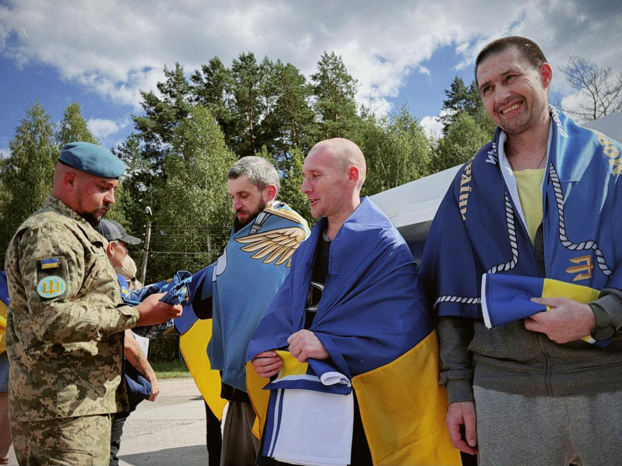 У межах спеціального етапу обміну через територію Білорусі, що відбувся 24 серпня, додому повернулися 10 українських захисників із лав ЗСУ та Нацгвардії, які вважалися зниклими безвісти на Донецькому напрямку. На фото двоє звільнених військовослужбовців праворуч щиро посміхаються, стоячи на рідній землі з накинутими на плечі державними прапорами України та символікою морської піхоти, тоді як захисник ліворуч залишається зосередженим, а представник Сил оборони у камуфляжній формі та береті допомагає їм із речами та прапорами.