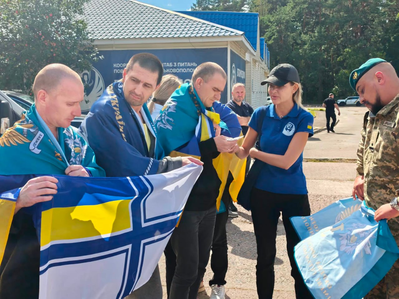 У межах спеціального етапу обміну через територію Білорусі, що відбувся 24 серпня, додому повернулися 10 українських захисників із лав ЗСУ та Нацгвардії, які вважалися зниклими безвісти на Донецькому напрямку. На фото звільнені військовослужбовці з накинутими на плечі державними прапорами України та символікою морської піхоти спілкуються з представницею та представником Координаційного штабу з питань поводження з військовополоненими на тлі будівлі хабу.