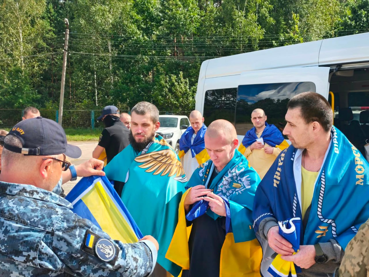 У межах спеціального етапу обміну через територію Білорусі, що відбувся 24 серпня, додому повернулися 10 українських захисників із лав ЗСУ та Нацгвардії, які вважалися зниклими безвісти на Донецькому напрямку. На фото звільнені військовослужбовці стоять біля білого пасажирського мікроавтобуса, загорнуті в державні прапори України та сині прапори морської піхоти, доки представники Сил оборони (один із них у піксельній формі Військово-морських сил) допомагають їм із речами та патріотичною символікою.
