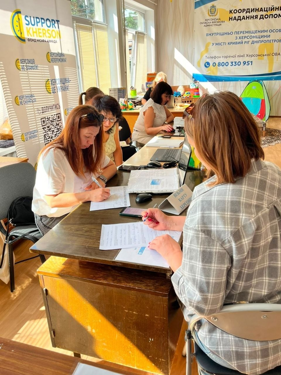 У Координаційному центрі «Вільні разом» у Кривому Розі у межах системної підтримки внутрішньо переміщених осіб продовжується реалізація проєкту «Картка Херсонщини». На фото відвідувачі та співробітники хабу працюють із документами й анкетами за довгими робочими столами, де встановлено інформаційну табличку «Видача Карти Херсонця». Реєстрація та видача карток відбуваються у світлому кабінеті на тлі офіційних банерів «SUPPORT KHERSON» та «Координаційний центр надання допомоги ВПО з Херсонщини у місті Кривий Ріг» із номером гарячої лінії Херсонської ОВА.