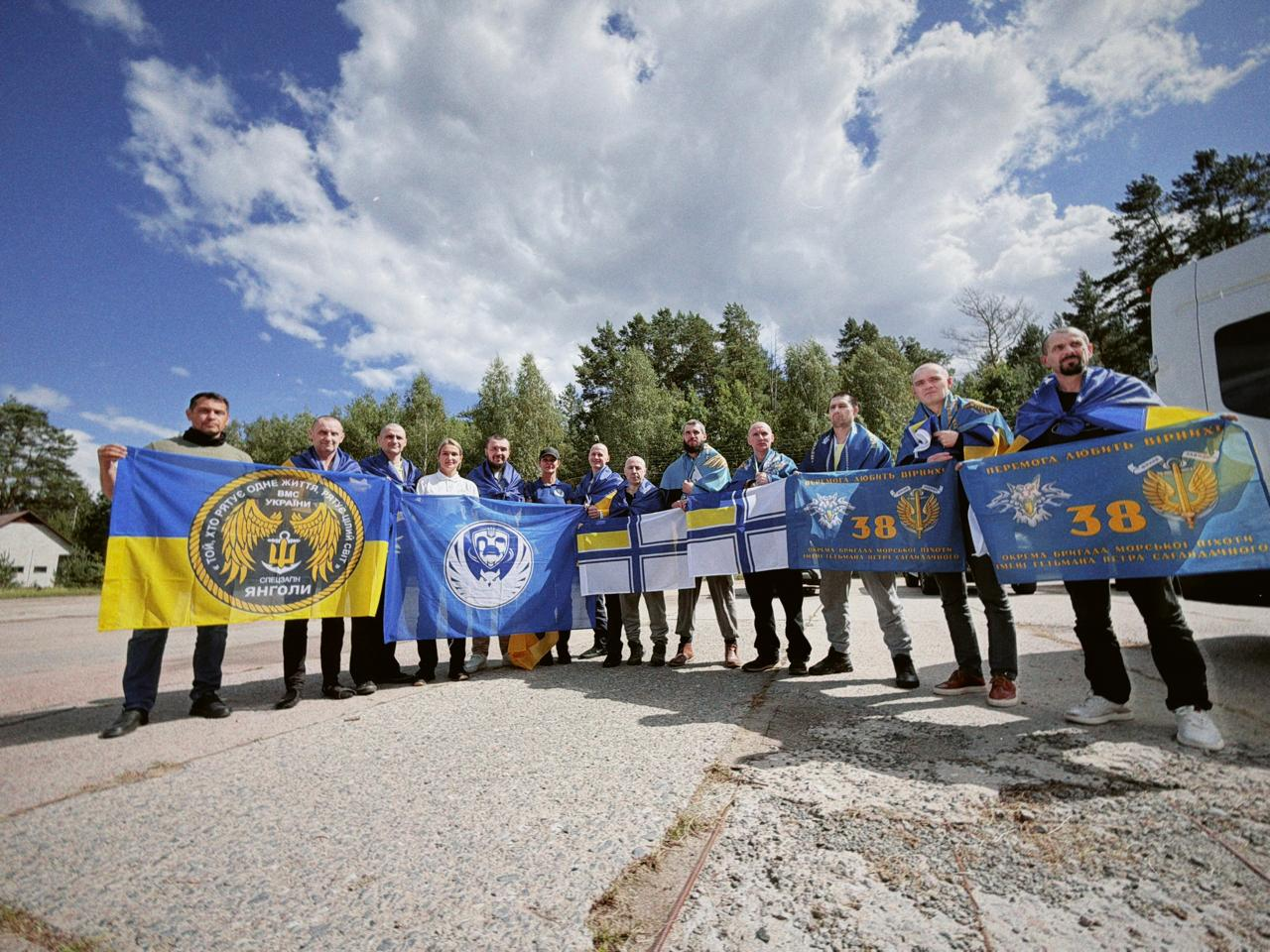 Група з десяти українських військових, серед яких 34-річний морпіх із Херсонщини, стоїть на дорозі після повернення з полону під час спеціального обміну 24 серпня 2026 року. Вони посміхаються на тлі синього неба та тримають прапори України, підрозділу ВМС «Янголи» та 38-ї окремої бригади морської піхоти.