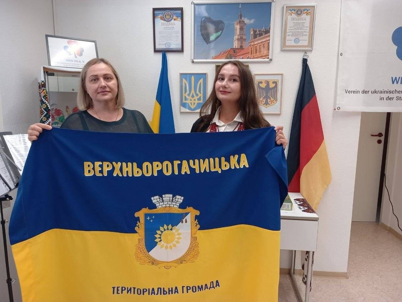 Представниці Верхньорогачицької громади Херсонщини святкують День Незалежності України в Німеччині. Жінка та дівчина у вишиванці посміхаються, тримаючи великий синьо-жовтий прапор своєї ТГ з офіційним гербом. Вони знаходяться у приміщенні німецько-українського товариства на тлі державних прапорів та гербів України й Німеччини, що символізує підтримку та збереження зв'язку з рідним краєм в евакуації.