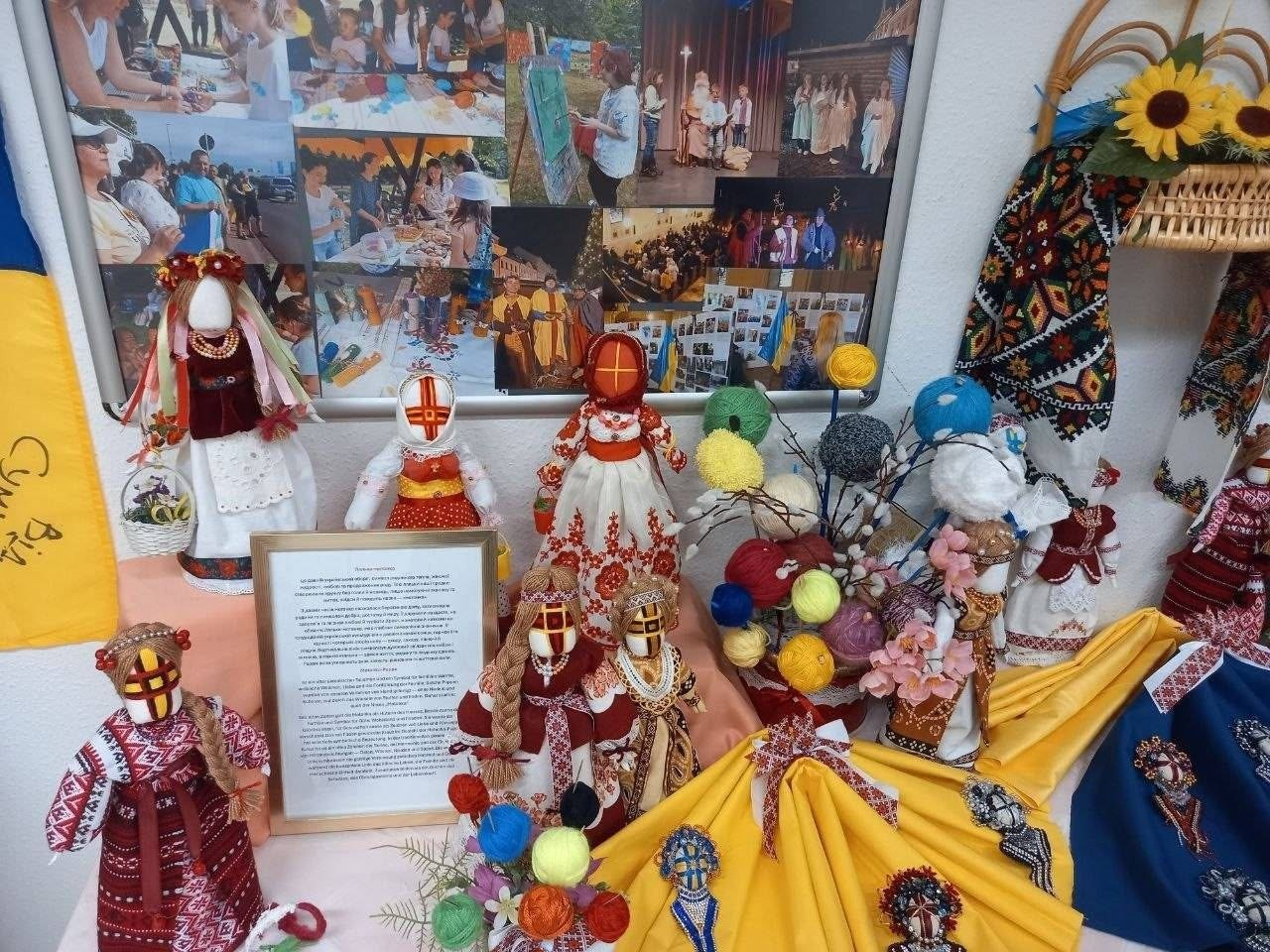 Виставка українських ляльок-мотанок, вишитих рушників та клубочків ниток, облаштована вихідцями з Верхньорогачицької громади в евакуації на святкуванні 35-ї річниці Незалежності України. Позаду виставки розміщено великий стенд із пам'ятними світлинами з культурних заходів.