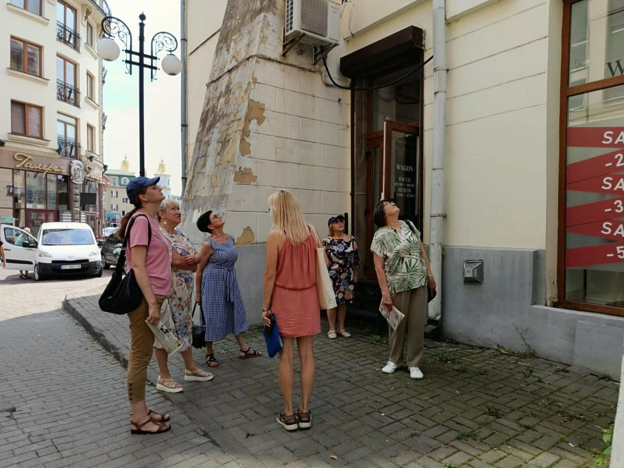 Пішохідна екскурсія «Вулицями Станіслава» для переселенок із Херсонщини в Івано-Франківську, організована центром «Вільні разом», Новомиколаївською СВА та Туристично-інформаційним центром для соціокультурної адаптації ВПО. У центрі кадру жінка-екскурсовод у рожевому комбінезоні показує вгору на архітектурні елементи старовинного будинку, а п'ять учасниць навколо неї зацікавлено розглядають фасад споруди. Тлом слугують затишна міська вулиця з бруківкою, кованим ліхтарем, вітринами крамниць та білим автомобілем неподалік.