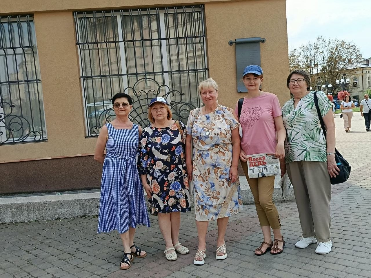Пішохідна екскурсія «Вулицями Станіслава» для переселенок із Херсонщини в Івано-Франківську, організована центром «Вільні разом», Новомиколаївською СВА та Туристично-інформаційним центром для соціокультурної адаптації ВПО. У центрі кадру п'ять учасниць заходу разом позують для колективного фото на бруківці під час прогулянки містом. Тлом слугує бежевий фасад будівлі з великими вікнами та декоративними кованими решітками, а одна з жінок тримає в руках свіжий випуск газети «Новий день».