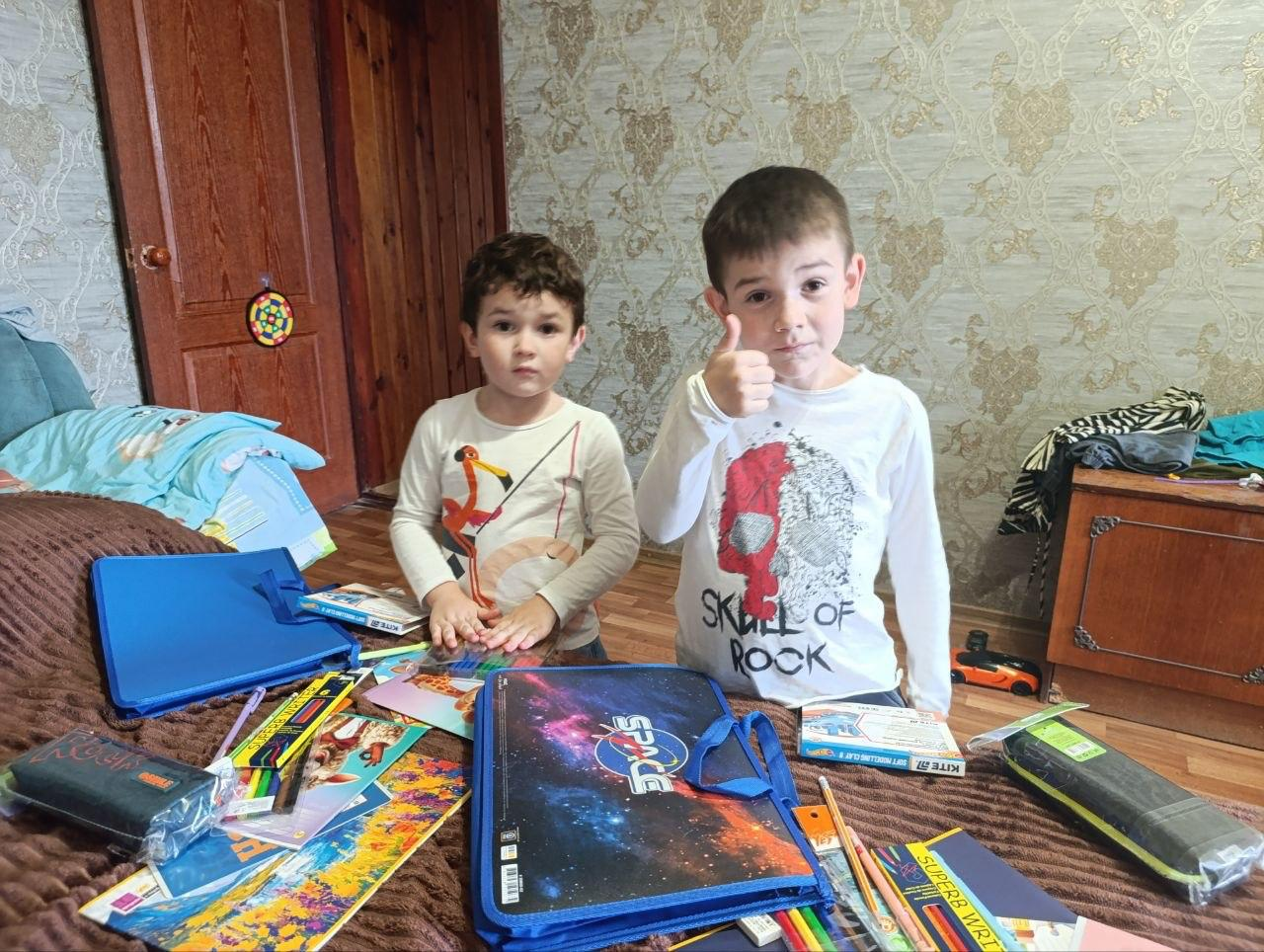Діти з Генічеської громади отримали канцелярські набори від Гайсинської громади Вінниччини напередодні нового навчального року у межах проєкту «Пліч-о-пліч: згуртовані громади». На передньому плані розкладено благодійну допомогу для юних переселенців: зошити, папки з космічним принтом та олівці марки «Kite». За речами стоять двоє хлопчиків-школярів, старший з яких радісно показує великий палець вгору на знак подяки партнерам за підтримку.
