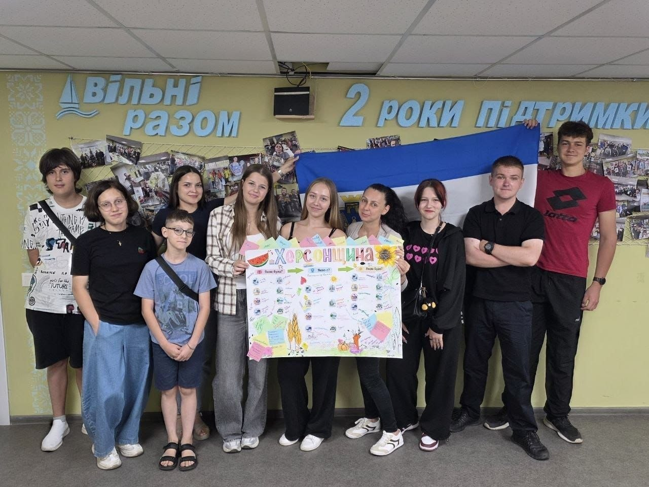 Учасники молодіжного флешмобу «Модель розвитку Херсонщини» позують для колективного фото у Координаційному центрі «Вільні разом» у Дніпрі. У центрі кадру група з десяти підлітків та молоді разом тримає великий створений ними плакат «Херсонщина» з малюнками й наліпками, а також розгорнутий синьо-білий прапор міста Херсон. Тлом слугує жовта стіна з великими об'ємними написами «Вільні разом» та «2 роки підтримки», на якій розвішано десятки виставкових фотографій.