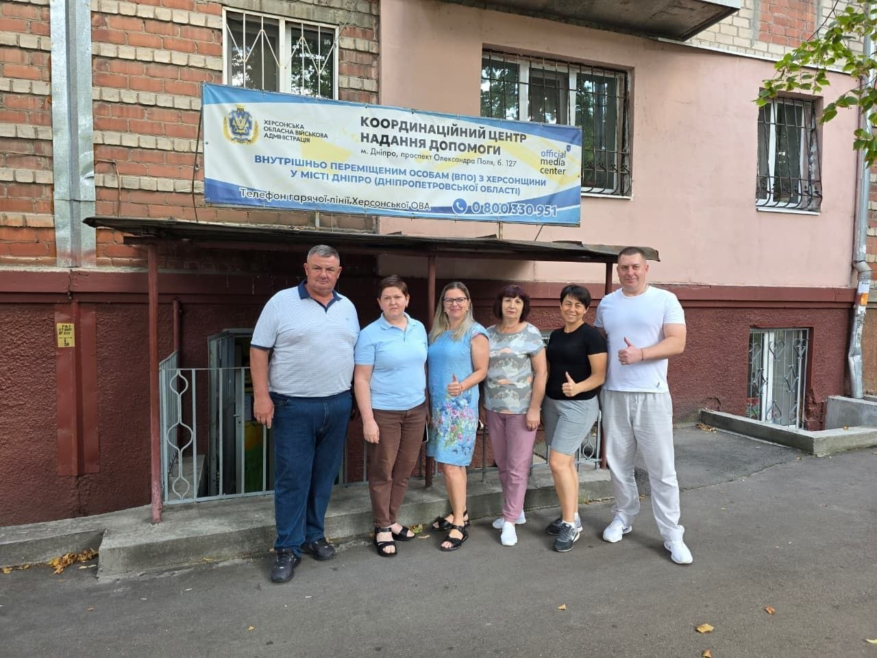 Зустріч партнерів біля Координаційного центру надання допомоги ВПО з Херсонщини у місті Дніпро в межах проєкту «Пліч-о-пліч: згуртовані громади». На фоні входу до центру стоїть група з шести осіб — четверо жінок і двоє чоловіків у літньому одязі, один з яких показує великий палець вгору. Над входом закріплено великий синьо-білий банер Херсонської ОВА з адресою хабу на проспекті Олександра Поля та номером гарячої лінії.