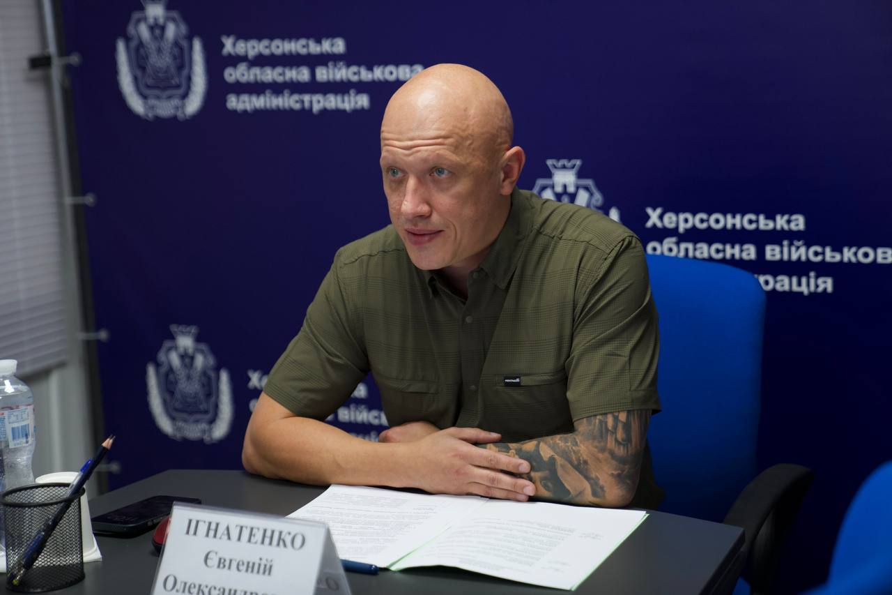 Заступник начальника Херсонської ОВА Євгеній Ігнатенко під час онлайн-зустрічі, присвяченої забезпеченню деокупованих громад області твердим паливом та зимовою підтримкою, де посадовець схилився над робочими документами та табличкою зі своїм ім'ям, сидячи на тлі синього офіційного банера з написами «Херсонська обласна військова адміністрація». Його зосереджений погляд спрямований убік екрана, де триває обговорення логістики допомоги для десятків тисяч домогосподарств регіону.