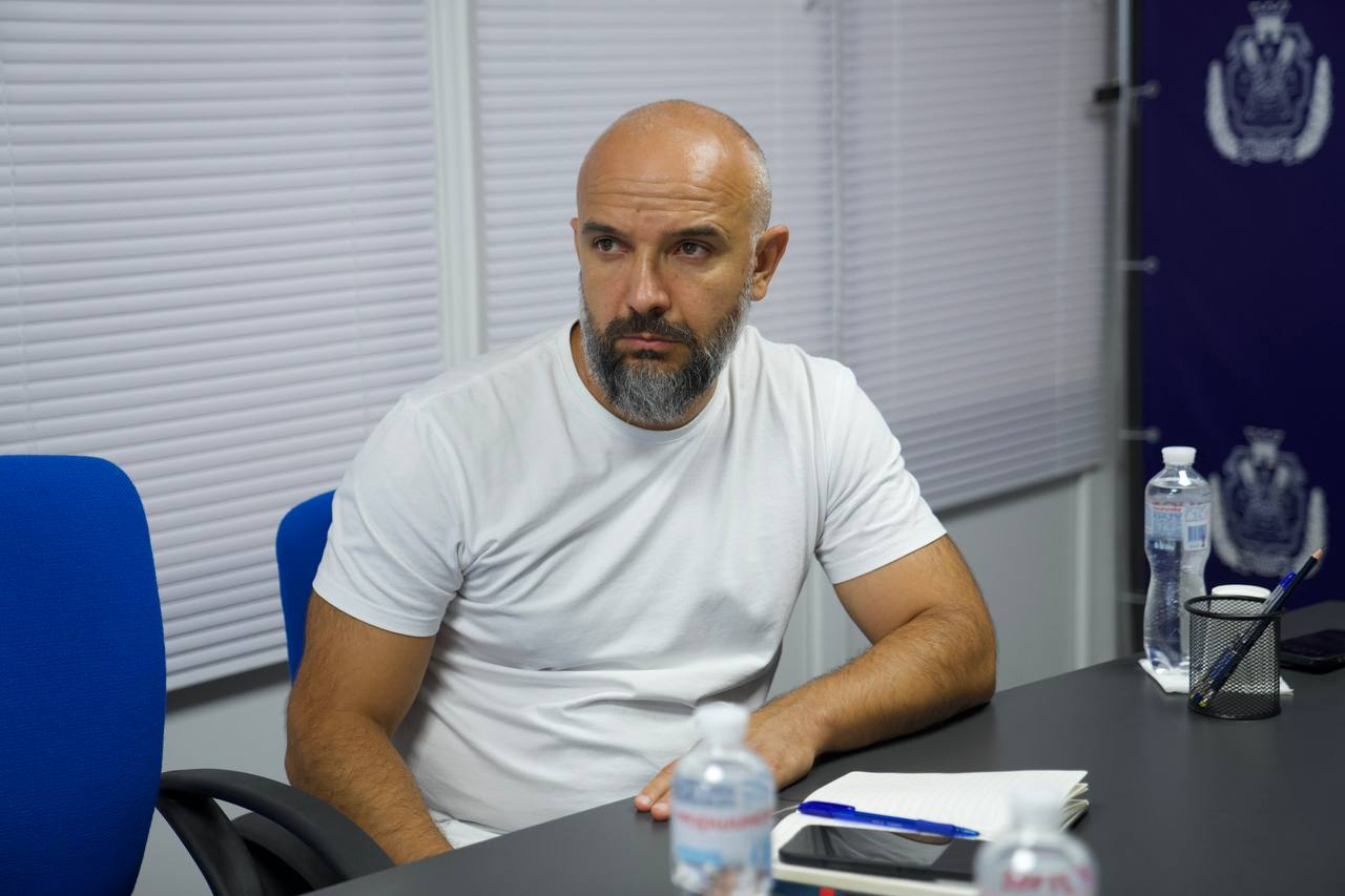 Заступник начальника Херсонської районної державної (військової) адміністрації Юліан Лучко бере участь у спільній робочій нараді щодо організації зимової підтримки та забезпечення твердим паливом десятків тисяч домогосподарств деокупованої Херсонщини. Посадовець зафіксований крупним планом за робочим столом в офіційному кабінеті на тлі білих жалюзі та синього банера з державною символікою, уважно слухаючи доповіді колег про координацію логістики та залучення міжнародних донорів.