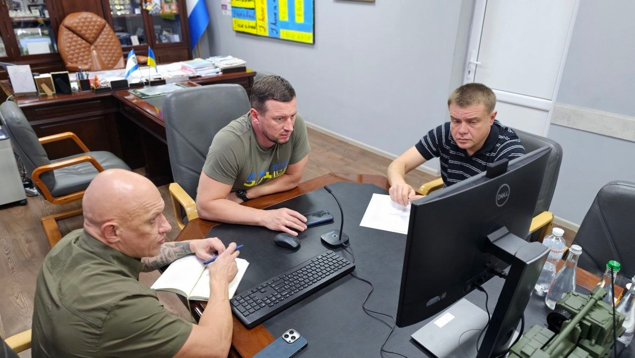 Начальник Херсонської ОВА Олександр Прокудін (по центру в зеленій футболці) проводить робочу нараду у службовому кабінеті разом із заступниками Євгенієм Ігнатенком (ліворуч) та Антоном Самойленком (праворуч на передньому плані) щодо ситуації з відновленням електропостачання та стану критичної інфраструктури, де керівники зосереджено аналізують дані на моніторі комп'ютера для координації роботи ремонтних бригад та безперебійного функціонування Пунктів Незламності.