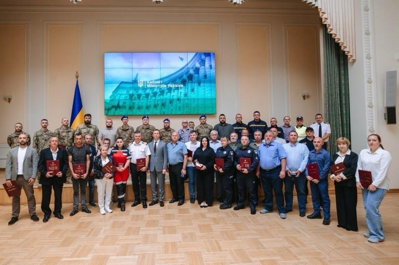 Групове фото з нагоди Дня Незалежності України, на якому у великій залі засідань на тлі прапора України та екрана з написом «Кабінет Міністрів України» зафіксовано нагородження працівників комунальної, енергетичної сфер та критичної інфраструктури Херсонщини. Понад тридцять відзначених фахівців — серед яких машиністка ТЕЦ Людмила Солонечна, менеджерка АЗС Любов Савченко, рятувальник ХОКАРС Олександр Довбиш та начальник теплоенергетичного району Анатолій Крохмаленко — стоять разом із колегами, військовими, поліцейськими та медиками, тримаючи в руках бордові папки з Подяками Прем’єр-міністра України.