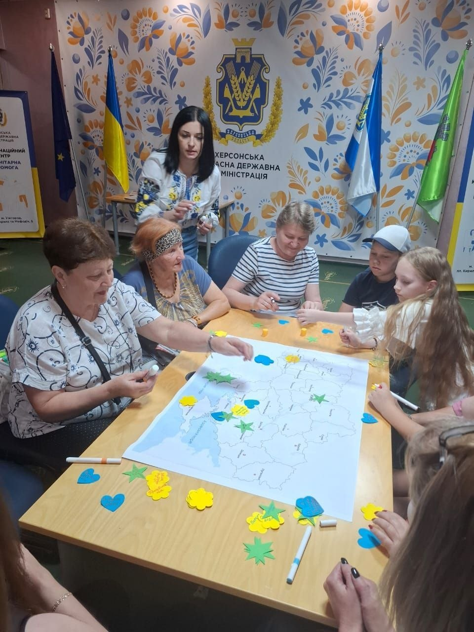 Група жінок та дітей у координаційному центрі «Вільні разом» в Ужгороді спільно створює патріотичний артоб'єкт «Карта Вдячності та Мрій» до 35-ї річниці Незалежності України. Учасники заходу сидять навколо столу та наклеюють паперові блакитно-жовті серця, зірки й квіти на карту, моделюючи майбутній розвиток Херсонщини під наглядом координаторки у вишиванці на тлі офіційного банера Херсонської ОДА.