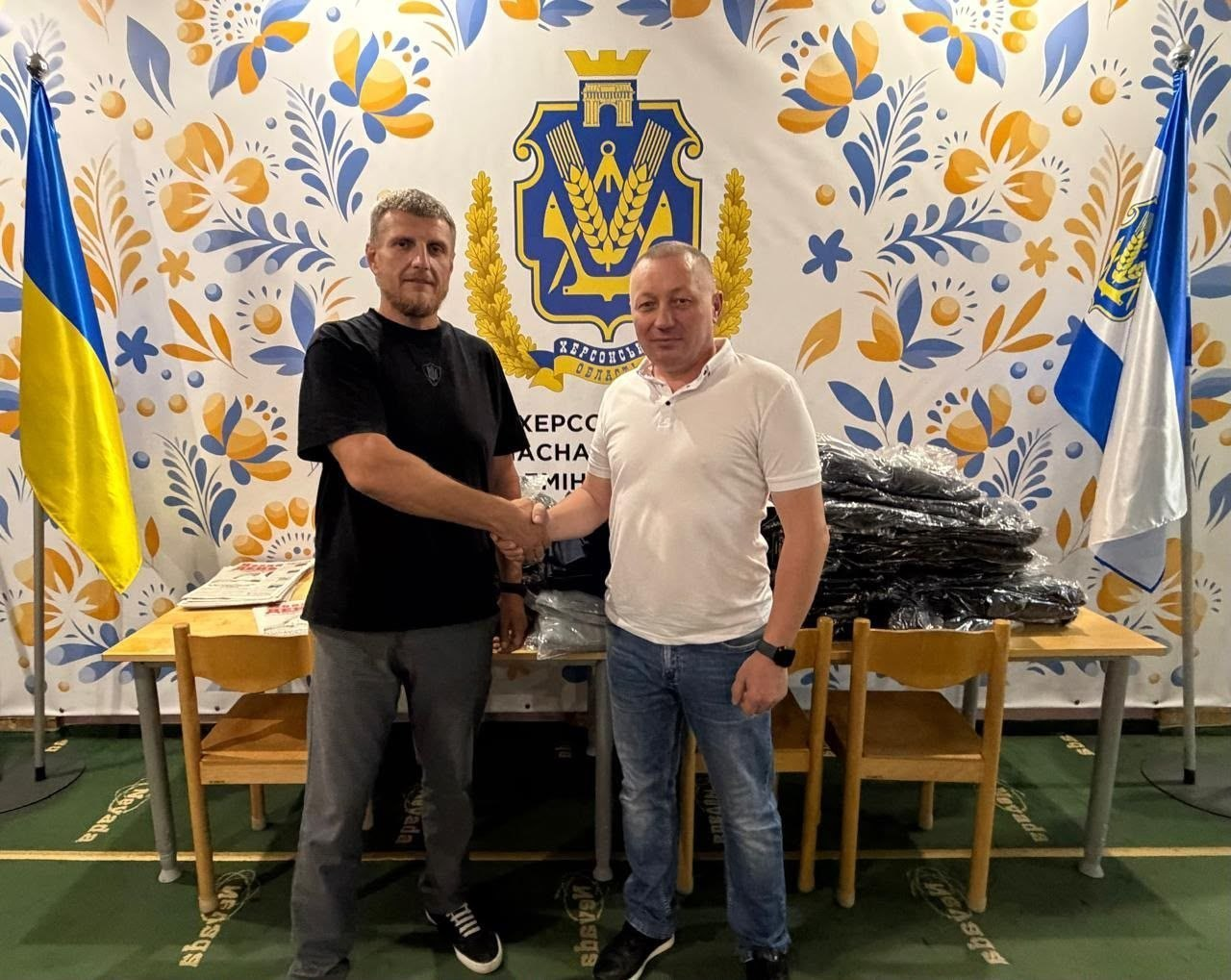 Начальник Нижньосірогозької СВА Віталій Кривчик (праворуч) та керівник координаційного центру «Вільні разом» в Ужгороді тиснуть один одному руки під час особистого прийому громадян та координації гуманітарної підтримки. Вони зафіксовані перед столом із пакунками допомоги на тлі офіційного банера з гербом Херсонської області, державним прапором України та прапором регіону.