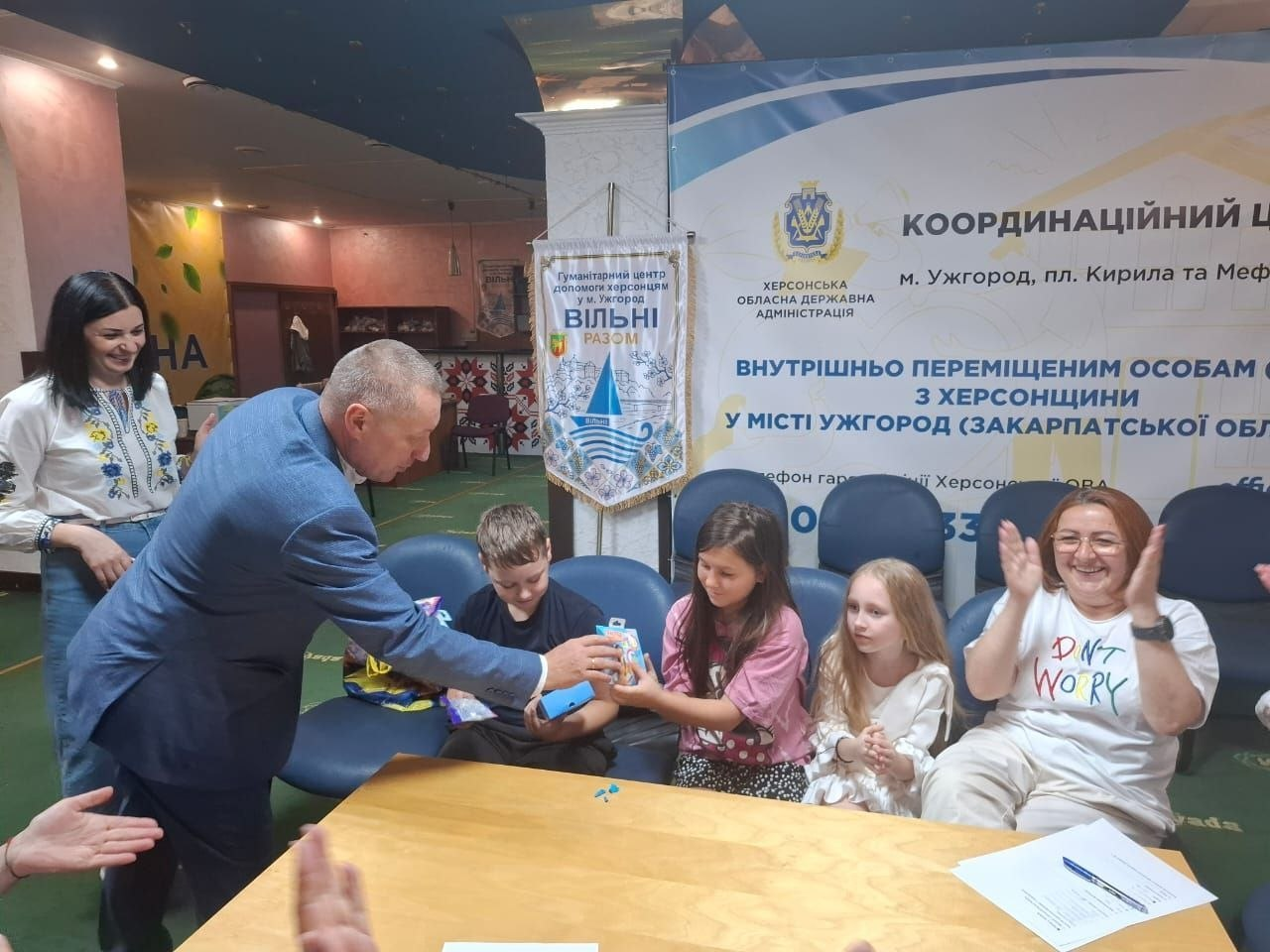 Начальник Нижньосірогозької СВА Віталій Кривчик під час особистого прийому громадян в ужгородському координаційному центрі «Вільні разом» передає подарунки та ігри дітям переселенців із Херсонщини. Керівник громади схилився біля столу, вручаючи пакунки хлопчику та дівчатам, за чим з усмішкою спостерігають матір дітей та координаторка хабу у вишиванці на тлі офіційного банера Херсонської ОДА.
