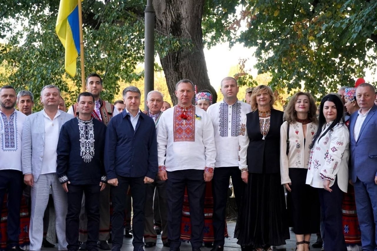 Групове фото посадовців Нижньосірогозької громади та представників Херсонщини на чолі з начальником Нижньосірогозької СВА Віталієм Кривчиком (крайній праворуч у синьому костюмі), які у вишиванках та святковому вбранні зафіксовані на вулиці під час мистецько-патріотичної акції «Молитва за Україну» в Ужгороді. Чоловіки та жінки стоять разом на тлі дерев і державного прапора України, об'єднавшись для вшанування пам'яті та покладання квітів.
