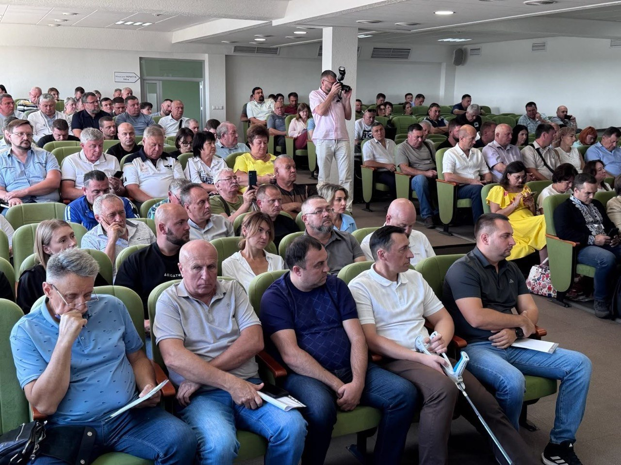 Заповнена аграріями та фермерами прифронтових областей велика зала засідань на Кіровоградщині під час наради з Міністром аграрної політики щодо викликів та підтримки сільгосппідприємств. Учасники заходу сидять у рядах зелених крісел амфітеатру, дехто тримає нотатки, а по центру між рядами стоїть фотограф, який документує зустріч.