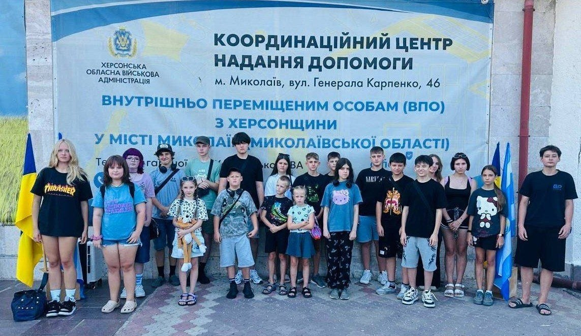 Група дітей та підлітків із родин ВПО з Чулаківської, Лазурненської, Білозерської, Бехтерської, Олешківської та Херсонської громад, які тимчасово проживають на Миколаївщині, зібралися біля гуманітарного хабу перед виїздом на оздоровлення та арттерапію до Волинської області. Діти стоять у ряд на тлі великого офіційного банера «Координаційний центр надання допомоги внутрішньо переміщеним особам з Херсонщини у місті Миколаїв та Миколаївській області» під егідою Херсонської ОВА. По боках від групи встановлені прапори України та міста Миколаєва. Поїздку організовано до Дня Незалежності України в межах 30-го туру благодійного проєкту «Теплі долоні» від ГО «Пятерня» та Департаменту соціального розвитку.
