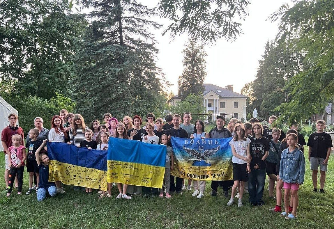 Група дітей та підлітків із родин ВПО з прифронтових громад Херсонщини разом із супроводжуючими позують на галявині біля Інтеграційного центру на Волині. Захід організовано Департаментом соціального розвитку Херсонської ОДА та ГО «Пятерня» в межах благодійного проєкту «Теплі долоні» до Дня Незалежності. Частина дітей вбрана у вишиванки, а на задньому плані серед дерев видніється світла будівля центру, де проходять заняття з арттерапії та психологічні тренінги.