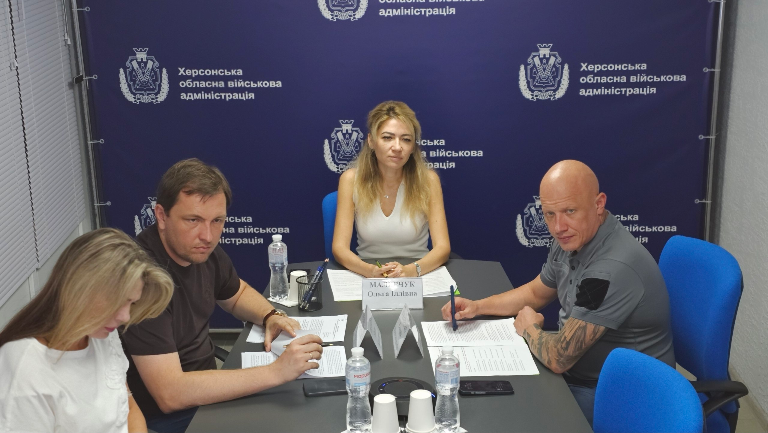 Світлина з робочої наради в Херсонській ОВА щодо захисту критичної інфраструктури деокупованих територій під головуванням заступниці начальника ХОВА Ольги Малярчук (у центрі кадру). Учасники наради сидять за сірим столом на тлі темно-синього брендованого банера з повторюваним написом «Херсонська обласна військова адміністрація». У центрі — Ольга Малярчук у бежевому топі, перед якою стоїть іменна табличка. Праворуч від неї сидить заступник начальника Херсонської ОВА Євгеній Ігнатенко, одягнений у сіре поло. Ліворуч за столом сидить директор Департаменту з питань цивільного захисту та оборонної роботи Євген Пісоцький у темній футболці, який уважно дивиться вперед. Поруч із ним схилилася над документами співробітниця профільного підрозділу. На столі розкладено робочі папери Комплексного плану стійкості області, стоять пляшки з водою, склянки та канцелярія.