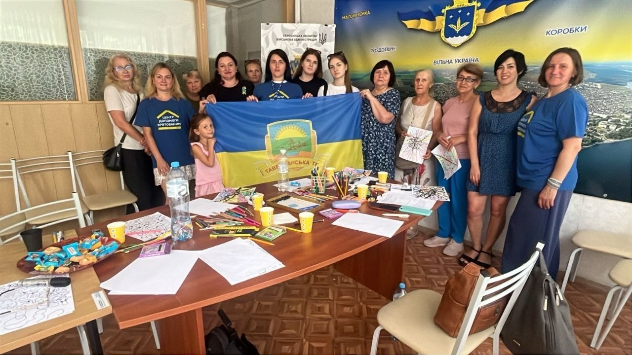 Група жінок та одна маленька дівчинка стоять у кімнаті навколо великого дерев'яного столу та тримають синьо-жовтий прапор Таврійської громади під час виїзного прийому для ВПО з Херсонщини в Координаційному центрі «Вільні разом» у Києві. На столі розкладено кольорові олівці, фломастери, папери з малюнками, солодощі та стаканчики. На задньому плані видно великий настінний банер із зображенням карти, герба та написами «Вільна Україна» і «Коробки», а також вертикальний логотип Херсонської обласної військової адміністрації.