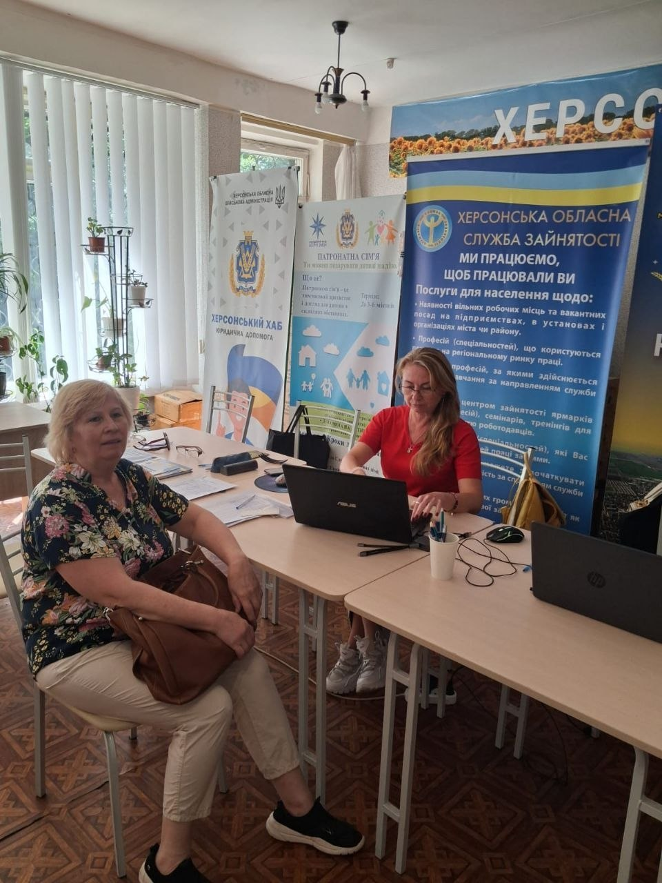 Світлина з профорієнтаційного тренінгу «50+ — час будувати нові кар'єрні плани» в Координаційному центрі «Вільні разом» у Києві для ВПО з Херсонщини.Фахівчиня Херсонського обласного центру зайнятості у червоній блузі сидить за столом з ноутбуком і консультує жінку щодо працевлаштування та грантової підтримки мікробізнесу.Поруч встановлено великий інформаційний банер із написом «Херсонська обласна служба зайнятості. Ми працюємо, щоб працювали ви».На задньому плані видно інші стенди, зокрема «Херсонський хаб. Юридична допомога» та плакат про патронатну сім'ю.