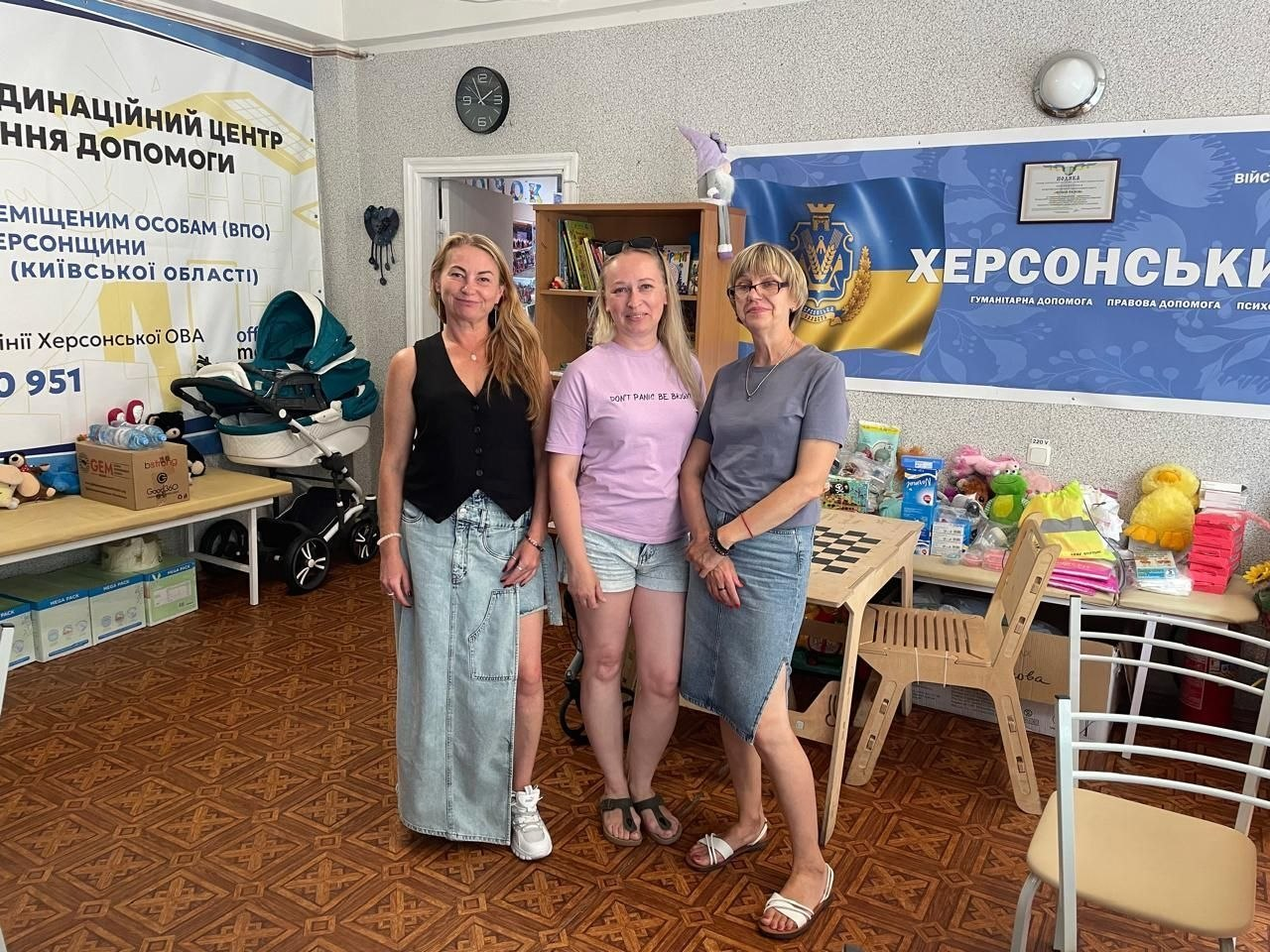 Три жінки стоять поруч у кімнаті Координаційного центру «Вільні разом» у Києві для підтримки ВПО з Херсонщини. На задньому плані ліворуч розташований великий інформаційний банер із написом «Координаційний центр надання допомоги внутрішньо переміщеним особам (ВПО) Херсонщини (Київської області)» та номером гарячої лінії. Праворуч на стіні висить темно-синій банер із гербом та великим написом «Херсонський хаб». У кімнаті також видно дитячі візочки, коробки з гуманітарною допомогою, іграшки та речі, розкладені на столах та полицях шафи.