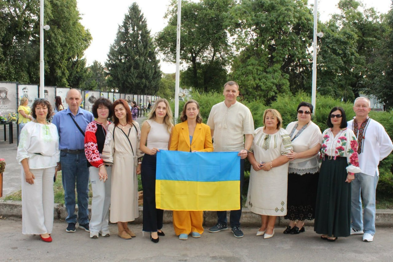 Групова світлина представників Козятинської, Генічеської та Ювілейної громад під час заходів до Дня Державного Прапора та Дня Незалежності України на Вінниччині в межах проєкту «Пліч-о-пліч: згуртовані громади». Усі одинадцять людей стоять на вулиці на тлі зелених дерев і паркових стендів. У центрі кадру начальник Генічеської міської військової адміністрації Євгеній Родіонов разом із міською головою Козятина Тетяною Єрмолаєвою в жовтому костюмі тримає розгорнутий синьо-жовтий прапор України. Ліворуч від них (п'ята зліва) стоїть представниця Ювілейної сільської громади Марина Ольшанська у світлій майці та темних штанах. Більшість інших учасників зустрічі одягнені в традиційні українські вишиванки, сукні з орнаментами та світлий одяг.