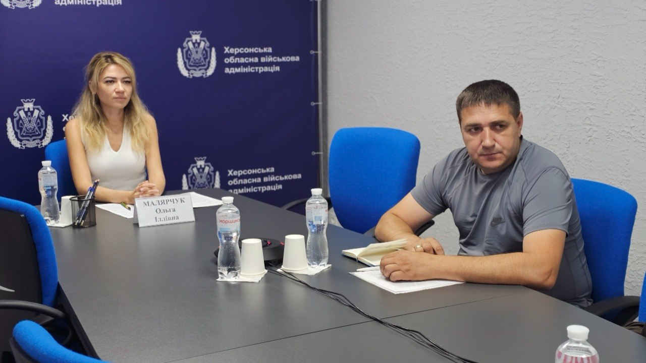 Світлина з онлайн-наради щодо вирішення проблемних питань осіб, зниклих безвісти за особливих обставин, та полонених Захисників і Захисниць України. За довгим сірим столом сидять двоє людей на тлі темно-синього брендованого банера з написом «Херсонська обласна військова адміністрація». Ліворуч сидить заступниця начальника Херсонської ОВА Ольга Малярчук у світлому топі. Праворуч за столом сидить заступник начальника управління взаємодії з правоохоронними органами Херсонської ОДА – начальник відділу координації роботи з правоохоронними органами Ігор Концевой у сірій футболці, який тримає руку біля відкритого робочого блокнота. Учасники наради обговорюють стан надання громадами Херсонщини фінансової підтримки дітям і членам сімей військовослужбовців та запуск цифрового помічника для ветеранів. На столі розкладено пляшки з водою, паперові стаканчики та канцелярію.