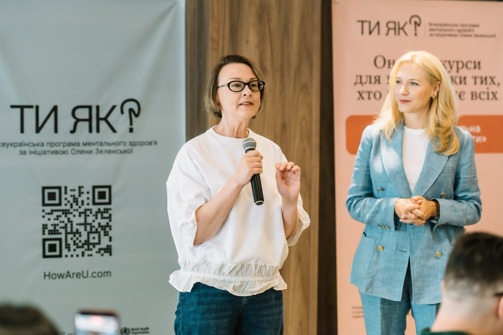 Світлина з виступу на навчальному заході для тренерів «Люди з досвідом війни. Гідність. Взаємодія: ветеранська політика, психоедукація та травмоінформований підхід» у межах «Національної платформи самоосвіти». У центрі кадру фахівчиня в окулярах та білій блузі виступає з мікрофоном у руках перед аудиторією регіональних амбасадорів Центрів зайнятості. Праворуч від неї стоїть колега у блакитному брючному костюмі. На задньому плані встановлено банери Всеукраїнської програми ментального здоров'я за ініціативою Олени Зеленської Ти як?.