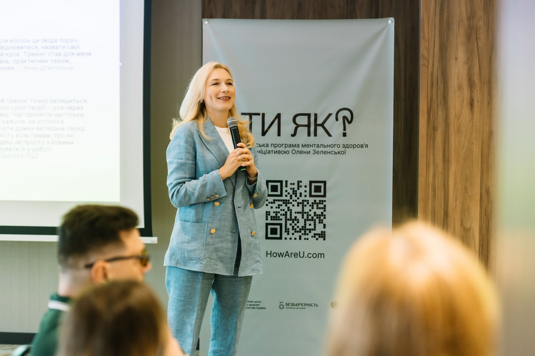 Гідність. Взаємодія: ветеранська політика, психоедукація та травмоінформований підхід» у межах Національної платформи самоосвіти. У центрі кадру спікерка в блакитному брючному костюмі виступає перед аудиторією регіональних амбасадорів Центрів зайнятості, тримаючи в руках мікрофон та посміхаючись. Праворуч на задньому плані розташований світлий банер Всеукраїнської програми ментального здоров'я з великим логотипом ТИ ЯК? за ініціативою Олени Зеленської, QR-кодом та адресою сайту HowAreU.com. Ліворуч на передньому плані видно профілі слухачів в аудиторії, а позаду спікерки — частину екрана проєктора з тезами виступу щодо практичних технік та психоедукації. Навчання спрямоване на підготовку фахівців Державної служби зайнятості до безбар'єрної роботи з ветеранами.