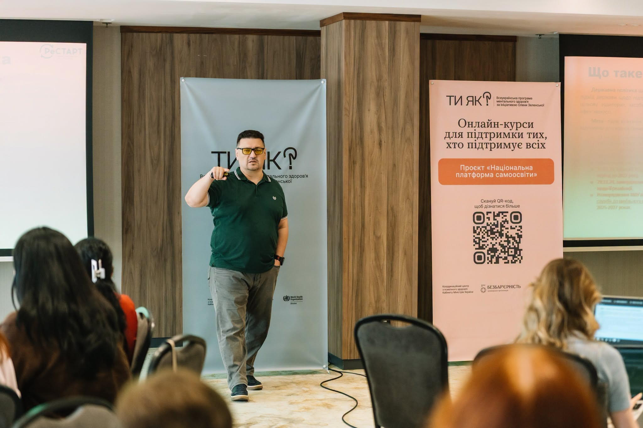 Світлина з виступу на навчальному заході для тренерів Люди з досвідом війни. Гідність. Взаємодія: ветеранська політика, психоедукація та травмоінформований підхід» у межах «Національної платформи самоосвіти. У центрі кадру спікер в окулярах, темно-зеленій футболці поло та сірих штанах виступає перед аудиторією регіональних амбасадорів Центрів зайнятості, піднявши руку під час промови. Праворуч від нього розташований рожевий інформаційний стенд із великим написом ТИ ЯК? Онлайн-курси для підтримки тих, хто підтримує всіх. Проєкт Національна платформа самоосвіти та великим QR-кодом. Ліворуч на задньому плані стоїть світлий банер Всеукраїнської програми ментального здоров'я Ти як?. На передньому плані видно потилиці слухачів в аудиторії, а з обох боків — частини великих екранів проєкторів. Навчання спрямоване на підготовку фахівців Державної служби зайнятості до безбар'єрної роботи з ветеранами.