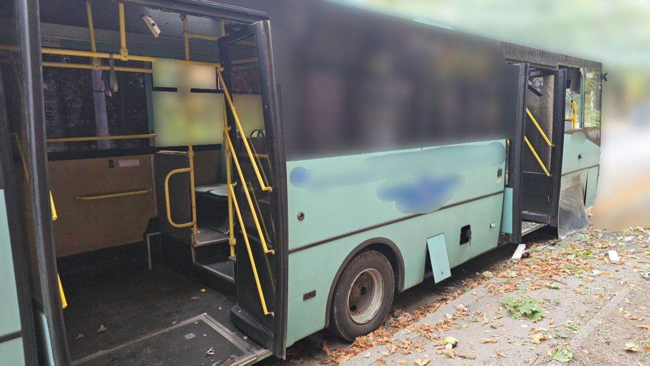 Міський автобус у Херсоні, який зазнав пошкоджень внаслідок атаки російського дрона. Транспортний засіб зелено-бірюзового кольору стоїть на узбіччі з відчиненими дверима, навколо на землі видно осіннє листя та дрібні уламки від вибуху. Салон автобуса порожній, видно жовті поручні, а верхня частина вікон та бортові написи розмиті.