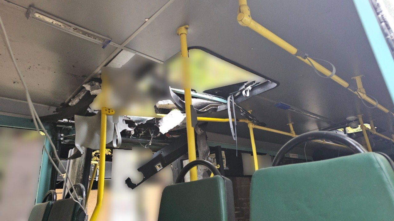 Вид ізсередини на понівечений салон міського автобуса в Херсоні після удару російського дрона. У стелі транспортного засобу утворилася велика наскрізна діра з рваними краями, з якої звисають шматки внутрішньої обшивки, утеплювача та пошкоджені дроти. На передньому плані видно спинки зелених пасажирських сидінь та вертикальні жовті поручні, а задня частина салону закрита розмиттям.