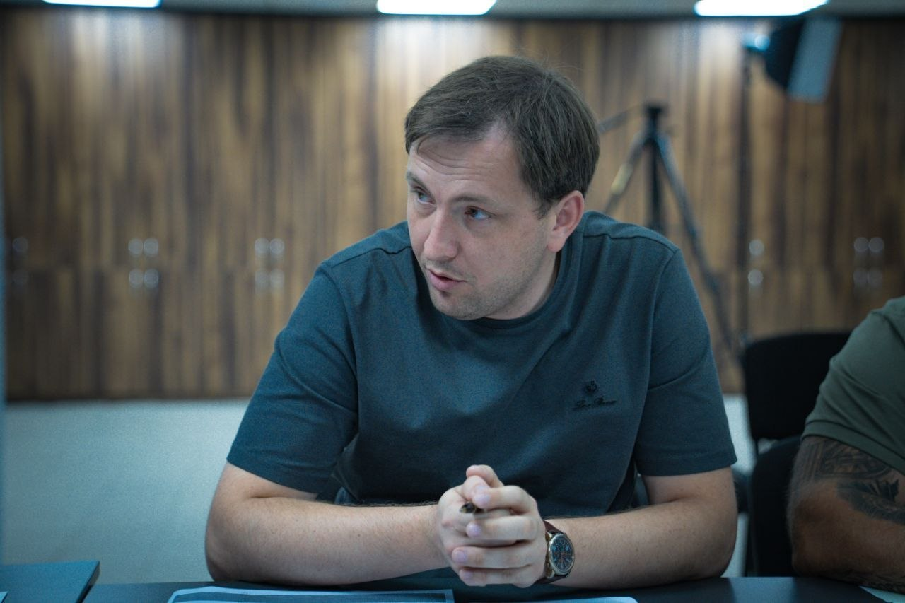 Директор Департаменту з питань цивільного захисту та оборонної роботи ХОВА Євген Пісоцький під час наради щодо захисту доріг області. Чоловік із коротким темним волоссям, одягнений у темно-сіру футболку та з наручним годинником, сидить за столом із зчепленими в замок пальцями та зосереджено слухає колег. Робоча зустріч проходить у кабінеті з вертикальними дерев'яними стіновими панелями.