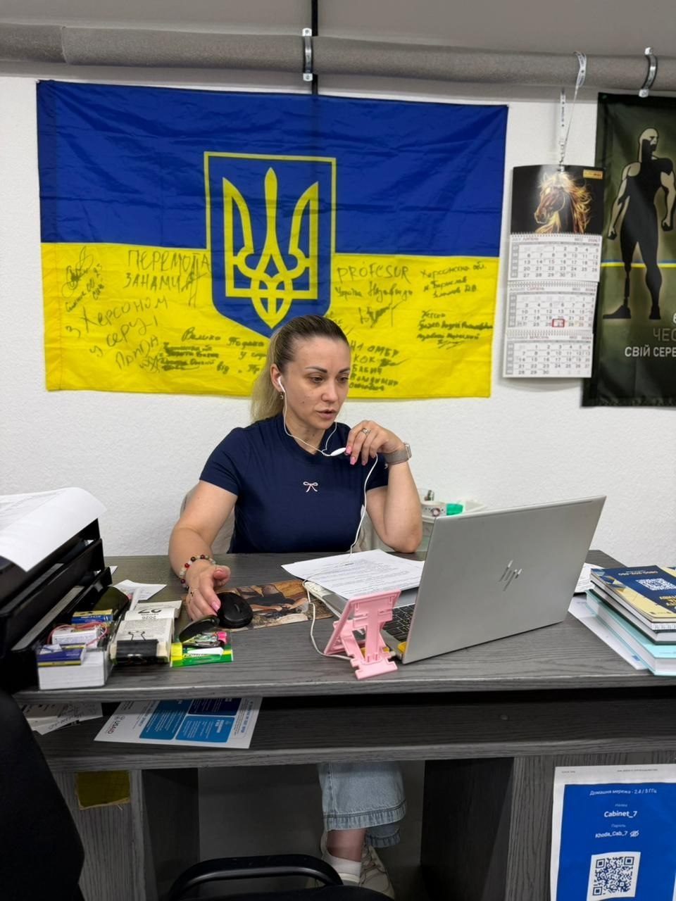 Начальниця управління у справах ветеранів Херсонської ОДА Дар’я Мазур за робочим столом під час онлайн-засідання обласної комісії. Посадовиця в темно-синій футболці та дротових навушниках зосереджено дивиться в екран ноутбука HP, опрацьовуючи документи щодо виплат родинам полеглих Захисників і Захисниць. На стіні за її спиною висить прапор України з великим золотим тризубом і підписами-побажаннями, а праворуч — настінний календар і плакат.