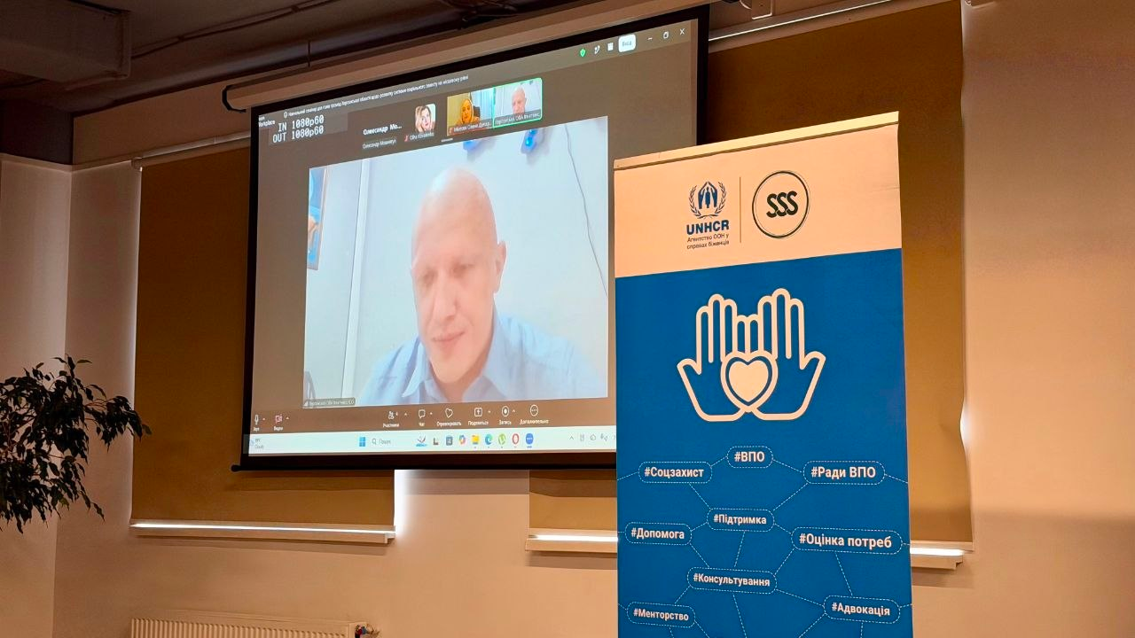 Зал проведення дводенного семінару для представників громад Херсонщини щодо розбудови місцевої системи соцзахисту. На великому проєкційному екрані транслюється онлайн-виступ заступника начальника Херсонської ОВА Євгенія Ігнатенка. Праворуч від екрана встановлено вертикальний банер із логотипами УВКБ ООН (UNHCR) та БО «Благодійний фонд “Стабілізейшен Суппорт Сервісез”» (SSS). На банері зображено емблему з долонями та серцем, а також наведено тематичні хештеги заходу: Соцзахист, ВПО, Ради ВПО, Підтримка, Допомога, Оцінка потреб, Консультування, Менторство та Адвокація.