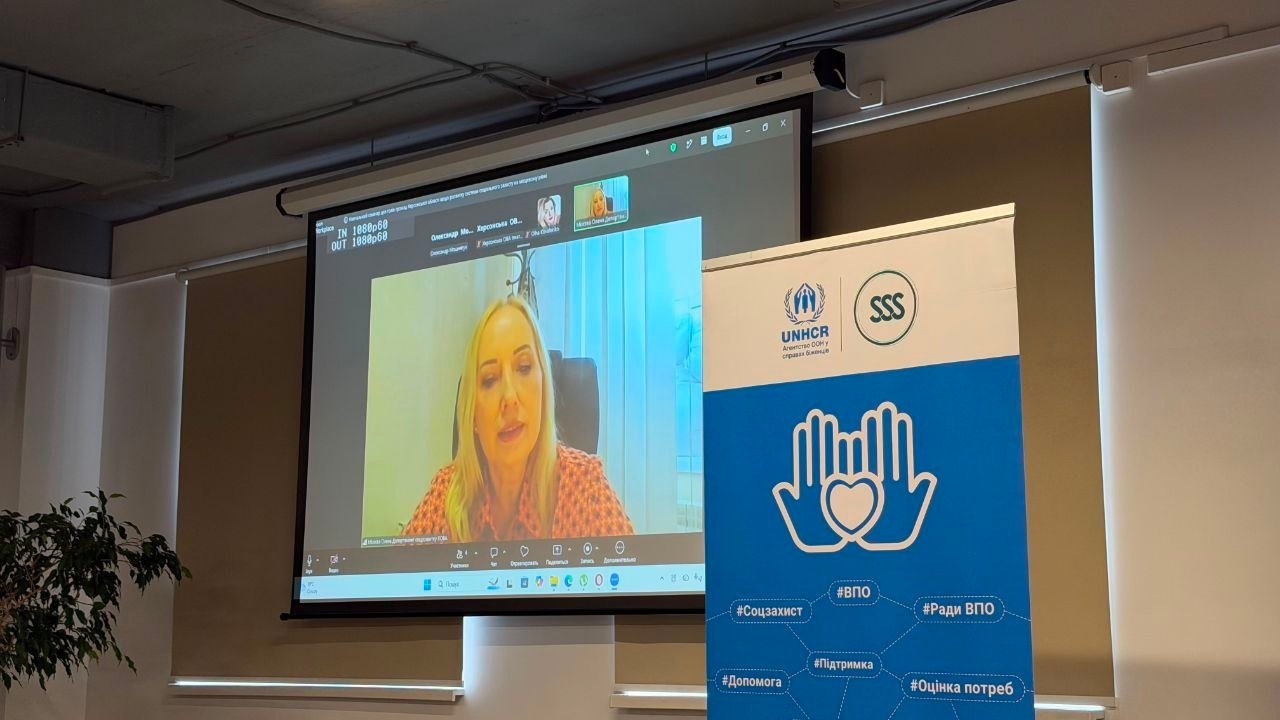 Зал проведення дводенного семінару для представників громад Херсонщини щодо розбудови місцевої системи соцзахисту. На великому проєкційному екрані по відеозв'язку транслюється онлайн-виступ директорки Департаменту соціального розвитку ХОДА Олени Міхеєвої. Світловолоса посадовиця в помаранчевому вбранні говорить до аудиторії через платформу відеоконференцій. Праворуч від екрана встановлено вертикальний банер із логотипами УВКБ ООН та БО «Благодійний фонд “Стабілізейшен Суппорт Сервісез”» (SSS) із синьою емблемою долонь та серця й тематичними хештегами.