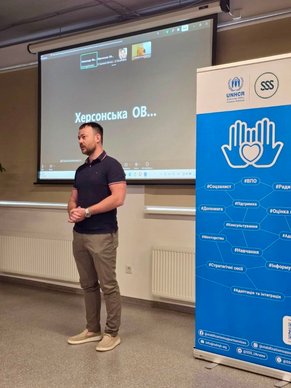 Керівник Агенції регіонального розвитку «Офіс євроінтеграції» Херсонської області Олег Кулакевич виступає на відкритті дводенного семінару з розбудови місцевої системи соцзахисту. Чоловік із короткою темною борідкою у чорній футболці поло та світло-коричневих штанях стоїть у залі біля великого проєкційного екрана, де відкрито онлайн-конференцію з написом «Херсонська ОВ...». Праворуч від нього встановлено інформаційний банер із логотипами УВКБ ООН та БО «Благодійний фонд “Стабілізейшен Суппорт Сервісез”» (SSS), прикрашений синьою графікою долонь із серцем та тематичними хештегами.