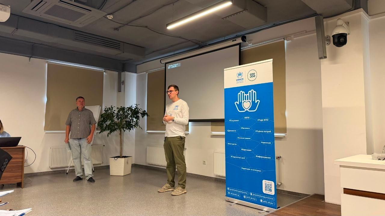 Один із тренерів виступає перед аудиторією під час дводенного навчального семінару для представників громад Херсонщини щодо розбудови місцевої системи соціального захисту. Чоловік в окулярах, білому лонгсліві з логотипом УВКБ ООН (UNHCR) та зелених штанях стоїть по центру залу біля великого білого проєкційного екрана. Ліворуч на задньому плані, біля фліпчарта та вазона з деревом, стоїть інший учасник заходу в сорочці в клітинку та джинсах. Праворуч на передньому плані встановлено вертикальний інформаційний банер із логотипами UNHCR та БО «Благодійний фонд “Стабілізейшен Суппорт Сервісез”» (SSS) із синьою емблемою долонь та серця й переліком хештегів.