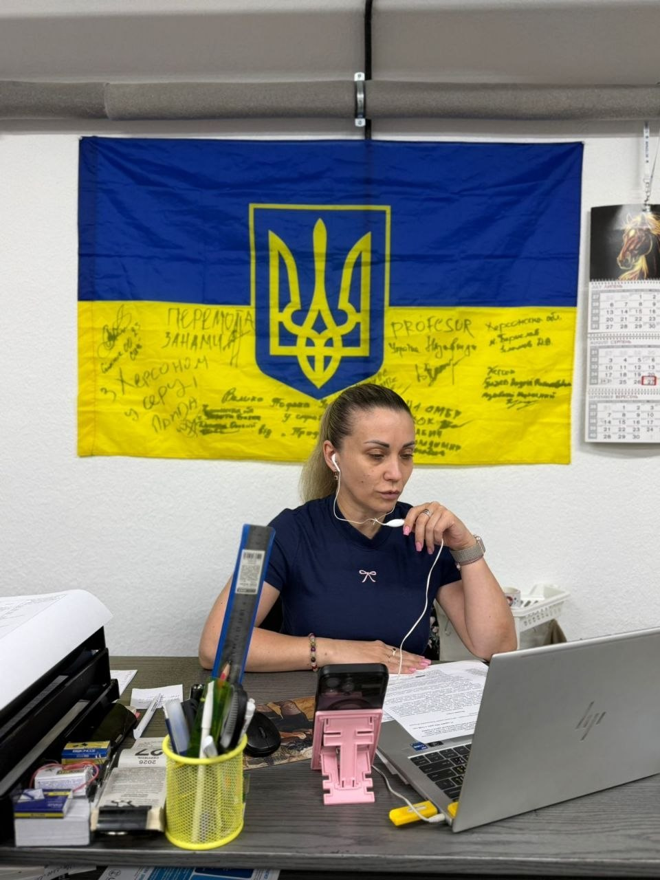 Начальниця управління у справах ветеранів Херсонської ОДА Дар’я Мазур за робочим столом під час онлайн-засідання обласної комісії щодо виплат звільненим із російського полону воїнам. Посадовиця в темно-синій футболці та білих дротових навушниках зосереджено дивиться в екран ноутбука HP, тримаючи пальцями мікрофон на дроті. На столі перед нею лежать документи щодо розгляду 11 заяв оборонців, стоїть рожевий тримач для телефона та жовта склянка з канцелярією. На стіні за її спиною висить Державний Прапор України з тризубом та підписами-побажаннями, а праворуч — настінний календар на серпень 2026 року.