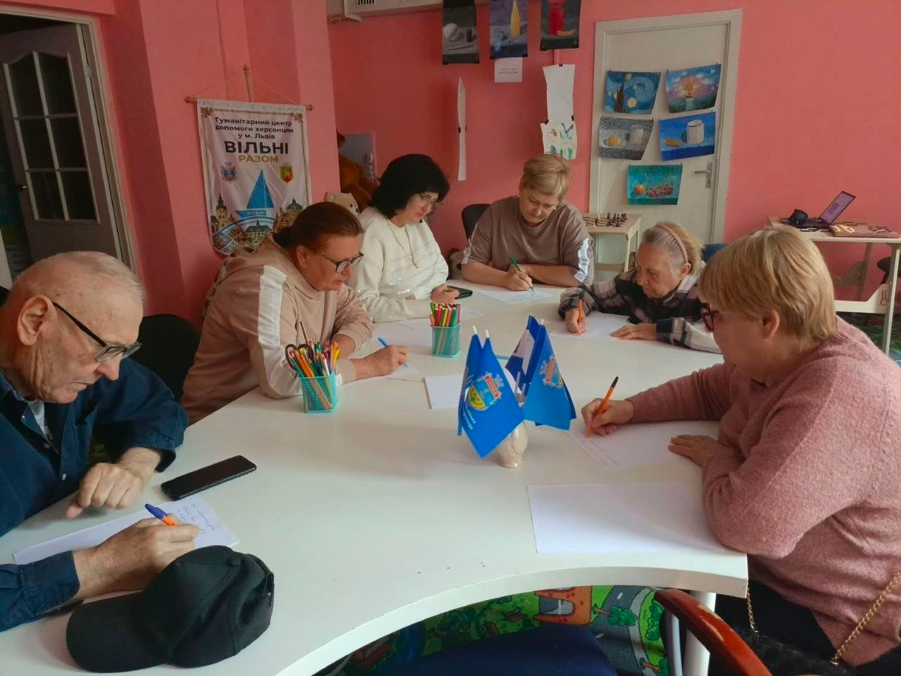 Переселенці з Херсонщини старшого віку під час оформлення та отримання «Картки Херсонщини» у львівському центрі «Вільні разом». Шість внутрішньо переміщених осіб (п'ять жінок та один чоловік ліворуч на передньому плані) сидять за великим білим столом і зосереджено заповнюють документи та роблять нотатки. Перед ними на столі лежать смартфони, необхідні для налаштування послуг, стоять підставки з олівцями та два маленькі сині прапорці. На задньому плані кімнати з рожевими стінами розміщено банер із написом «Гуманітарний центр допомоги херсонцям у м. Львів “Вільні разом”».