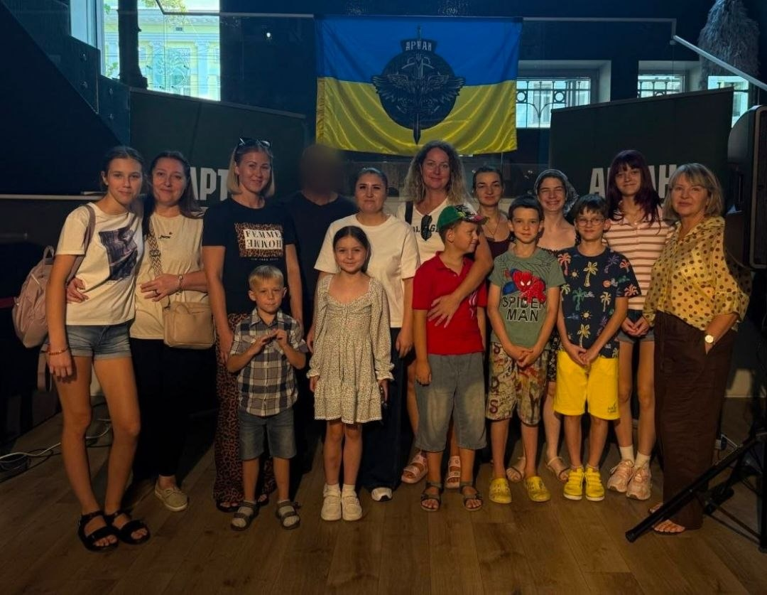 Переселенці з Херсонщини під час візиту до відкритого простору «Артан» в Одесі. Велика група дорослих і дітей позує для спільного фото в затемненому залі з дерев'яною підлогою. Захід, присвячений Дню пам’яті захисників України, організувала служба у справах дітей Верхньорогачицької селищної ради. На стіні позаду учасників висить прапор України з великим темним шевроном спецпідрозділу «Артан» по центру. Обличчя військовослужбовця на задньому плані розмите з міркувань безпеки. Діти та дорослі зафіксовані після ознайомлення з бойовим шляхом підрозділу та випробування симуляторів дронів.